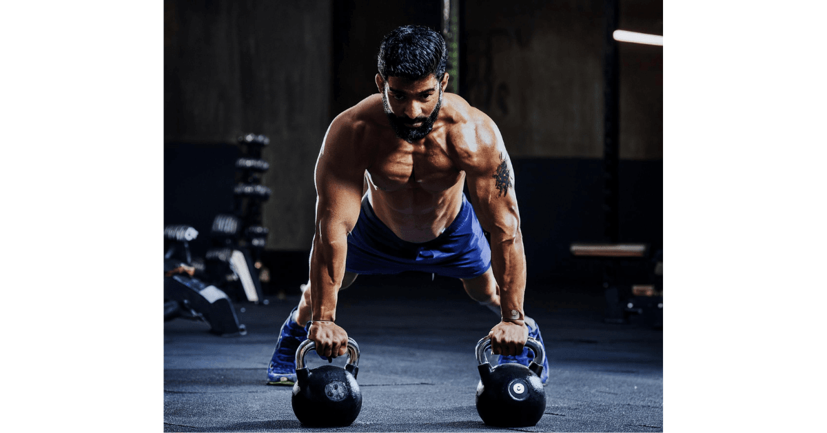 Shirtless man performing a kettlebell push-up exercise in a gym, showcasing strong chest and arm muscles while holding two kettlebells, focused on his worko
