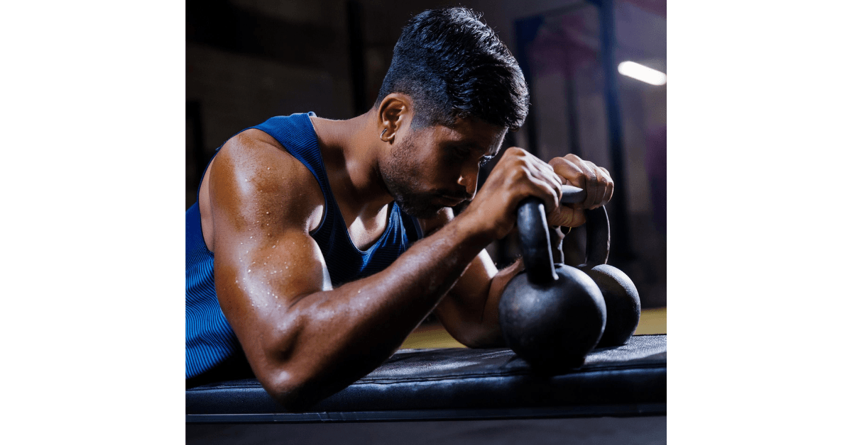 Close-up of a man in a blue tank top, sweating and resting his arms on kettlebells during a workout, highlighting his muscular arms and intense focus.